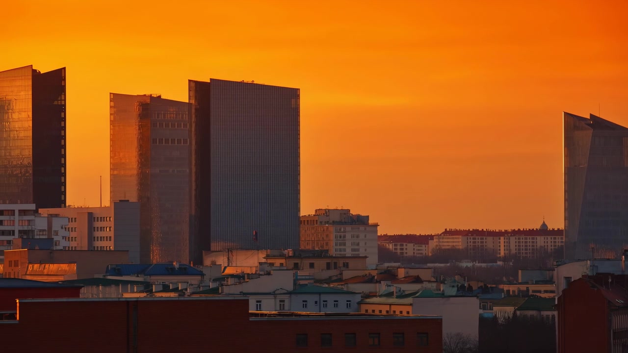 Movimento panorâmico lento revela torres de vidro agrupadas e telhados urbanos mais baixos brilhando na luz quente do entardecer, enquanto um céu laranja intenso preenche o fundo
