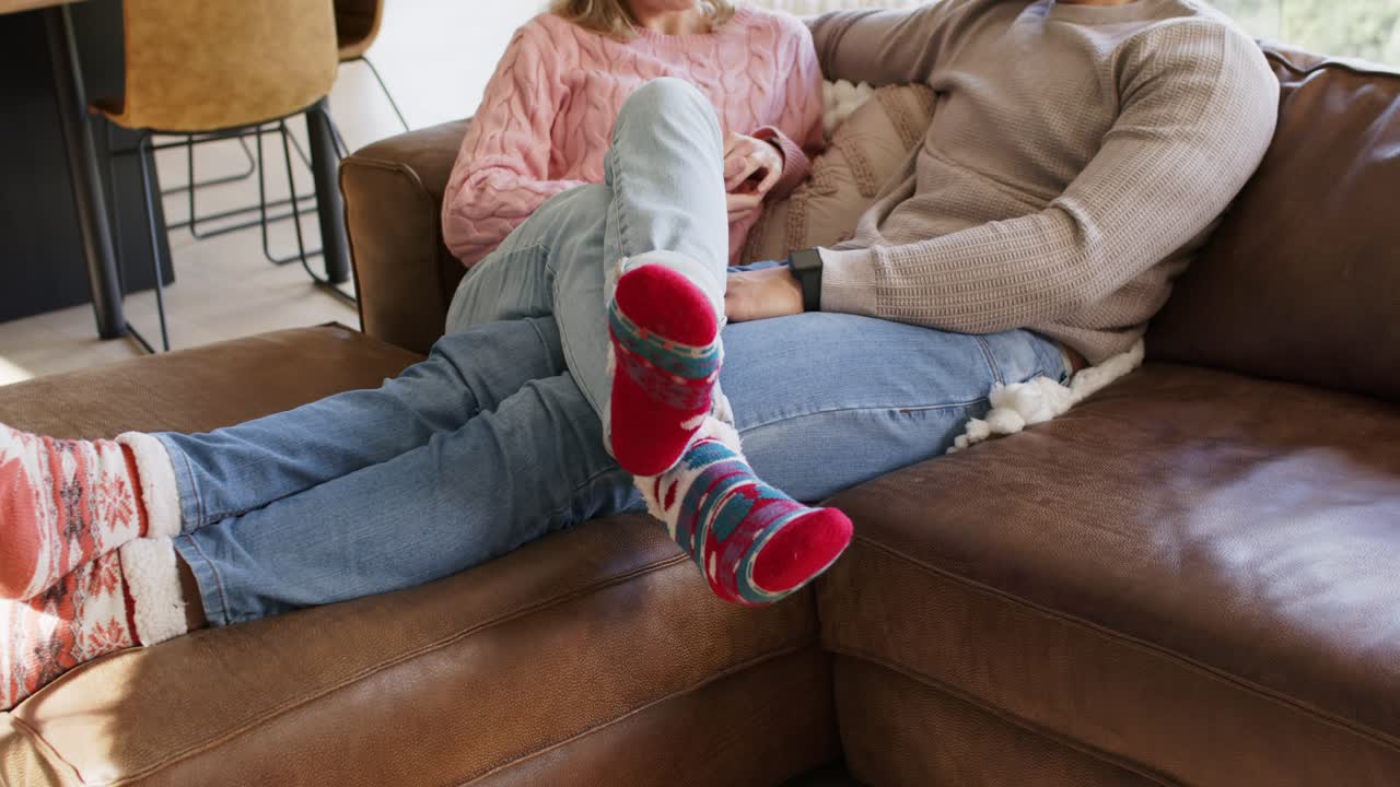 Couple settling on brown leather sofa to relax, woman swinging legs onto man's lap