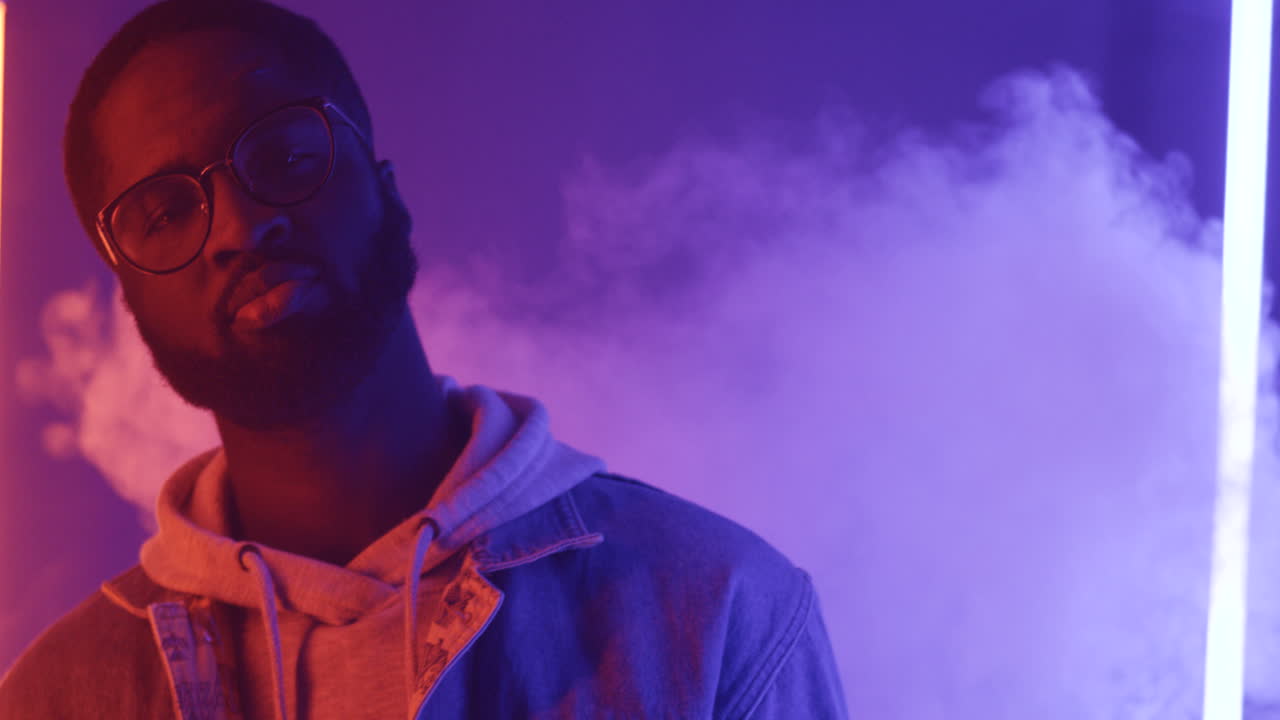 tampilan dekat pria bergaya amerika berkacamata menari memegang smartphone dan melihat kamera pada latar belakang cahaya neon merah muda dan ungu