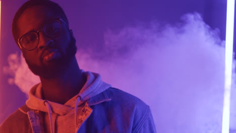 Close-Up-View-Of-American-Stylish-Man-In-Glasses-Dancing-Holding-Smartphone-And-Looking-At-Camera-On-Pink-And-Violet-Neon-Light-Background