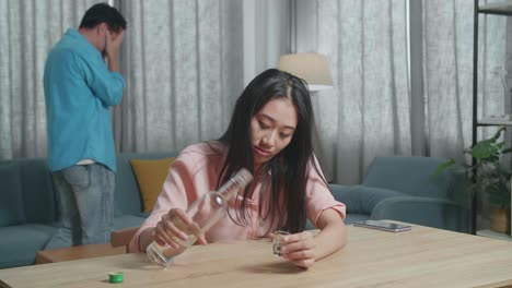 술에 취한 우울한 아시아 여성이 술을 마시기 전에 글라스에 보드카를 부어 울고, 집에서 그녀의 뒤를 따라 는 남자