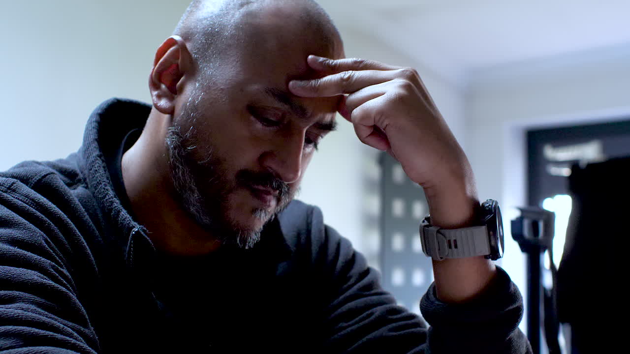 A man sits, his hand scratching his head, his expression revealing the burden of stress and anxiety he carries, vividly illustrating the tangible strain he experiences