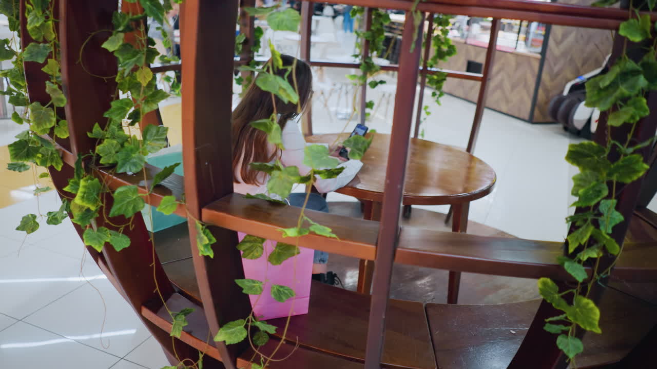 장식 된 나무 좌석에 앉아있는 멋진 젊은 여성, 전화를 들고, 그녀의 에 쇼핑 가방, 쇼핑몰의 바쁜 레스토랑 배경에서 움직이는 사람들과