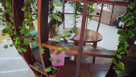 chic mujer joven sentada en un sentado de madera decorativa, sosteniendo el teléfono, bolsas de compras a su lado, con la gente moviéndose en el bullicioso fondo del restaurante del centro comercial
