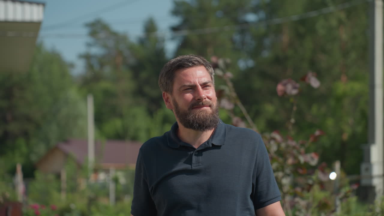 Man standing in sunlight on farm wiping forehead with hand while holding plier, light reflecting off sweaty head, looking into distance during inspection pause with trees and house in background