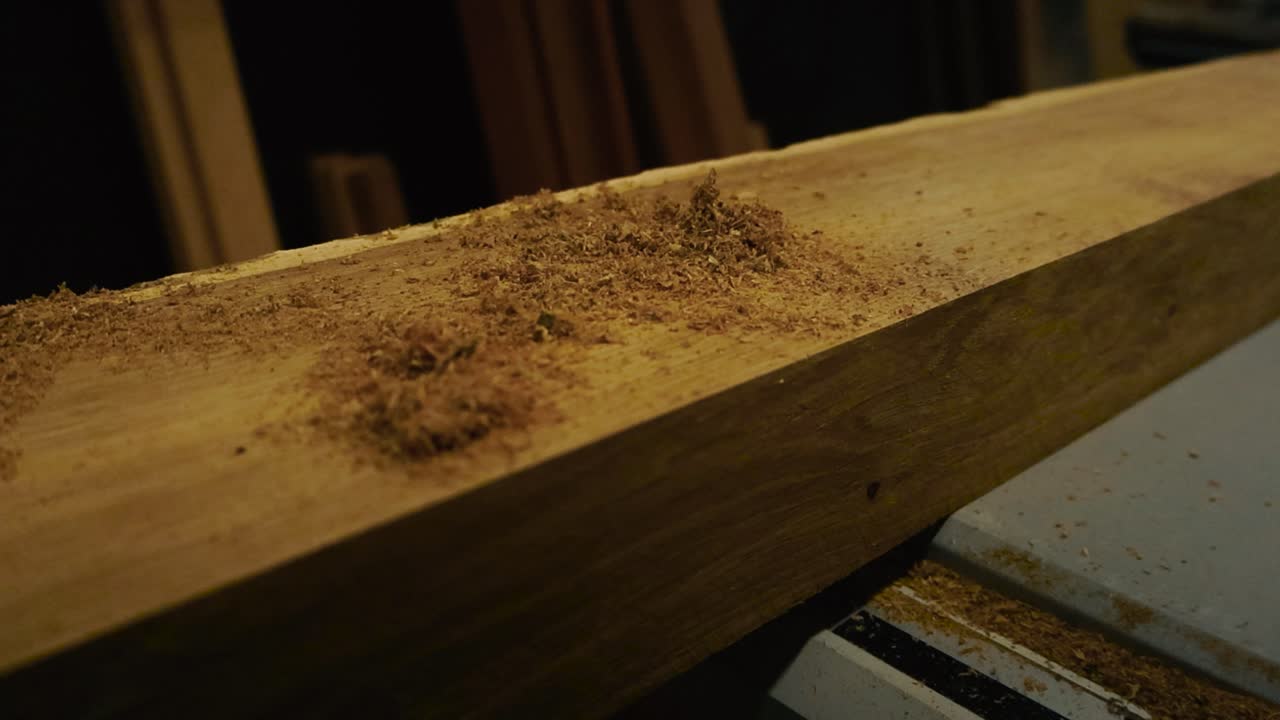 Sawdust and wood chips being blown off and away from a wooden oak yellow and brown colored slab that is on a table saw in a workshop indoors with bokeh blurry background. Flying in slow motion