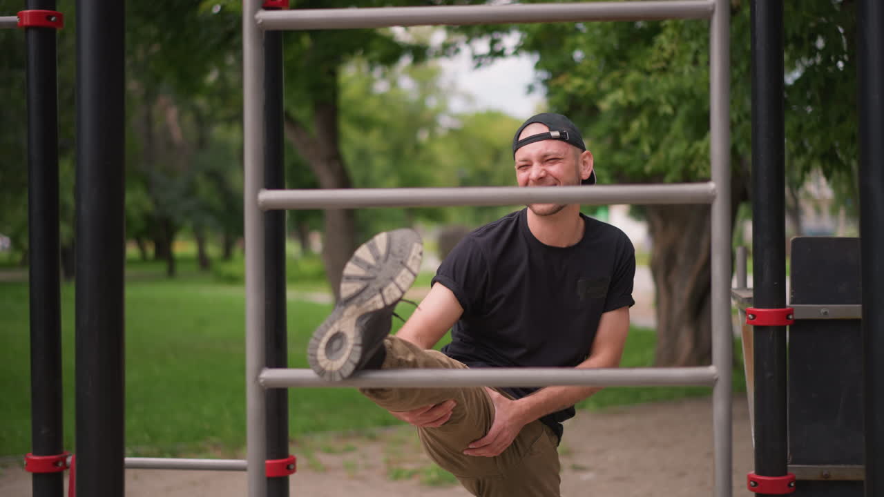 Park Mobility Leg Stretch On Metal Bars With White Male Smiling And Warming Up, Relaxed Atmosphere, Casual Cap And Cargo Pants, Focus On Hamstring Flexibility And Joint Range Of Motion