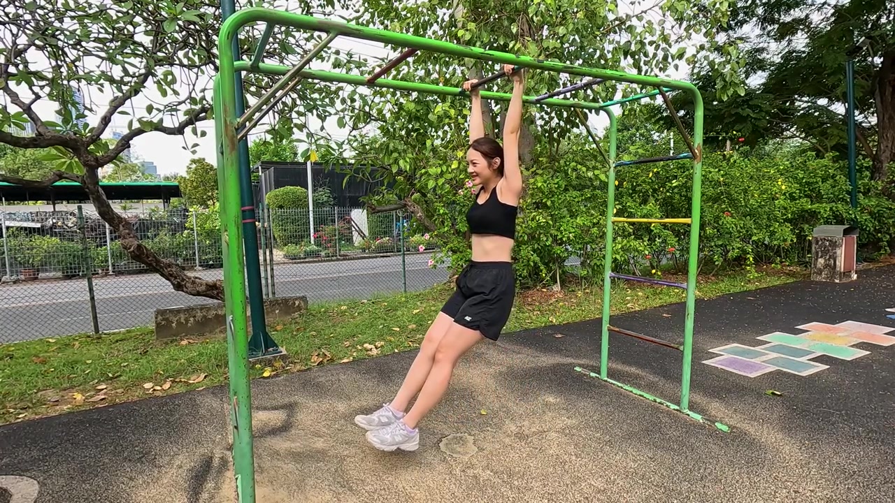 Fit Asian woman hangs from green monkey bars during outdoor workout in natural daylight.