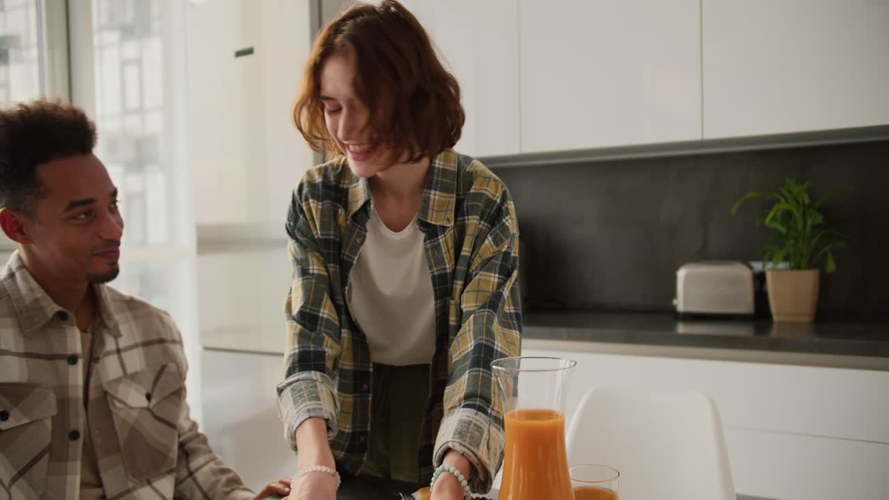 Happy young adult girl with brown hair and bob hairstyle serves fresh croissants to the table during breakfast with her young Black brunette boyfriend at the table in a modern apartment in the kitchen