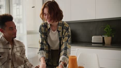 Happy-young-adult-girl-with-brown-hair-and-bob-hairstyle-serves-fresh-croissants-to-the-table-during-breakfast-with-her-young-Black-brunette-boyfriend-at-the-table-in-a-modern-apartment-in-the-kitchen