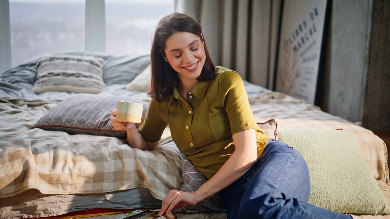 Nærbillede af en afslappet brunette, der drikker kaffe, mens hun sidder på tæppet i et solbeskinnet hjem om morgenen.