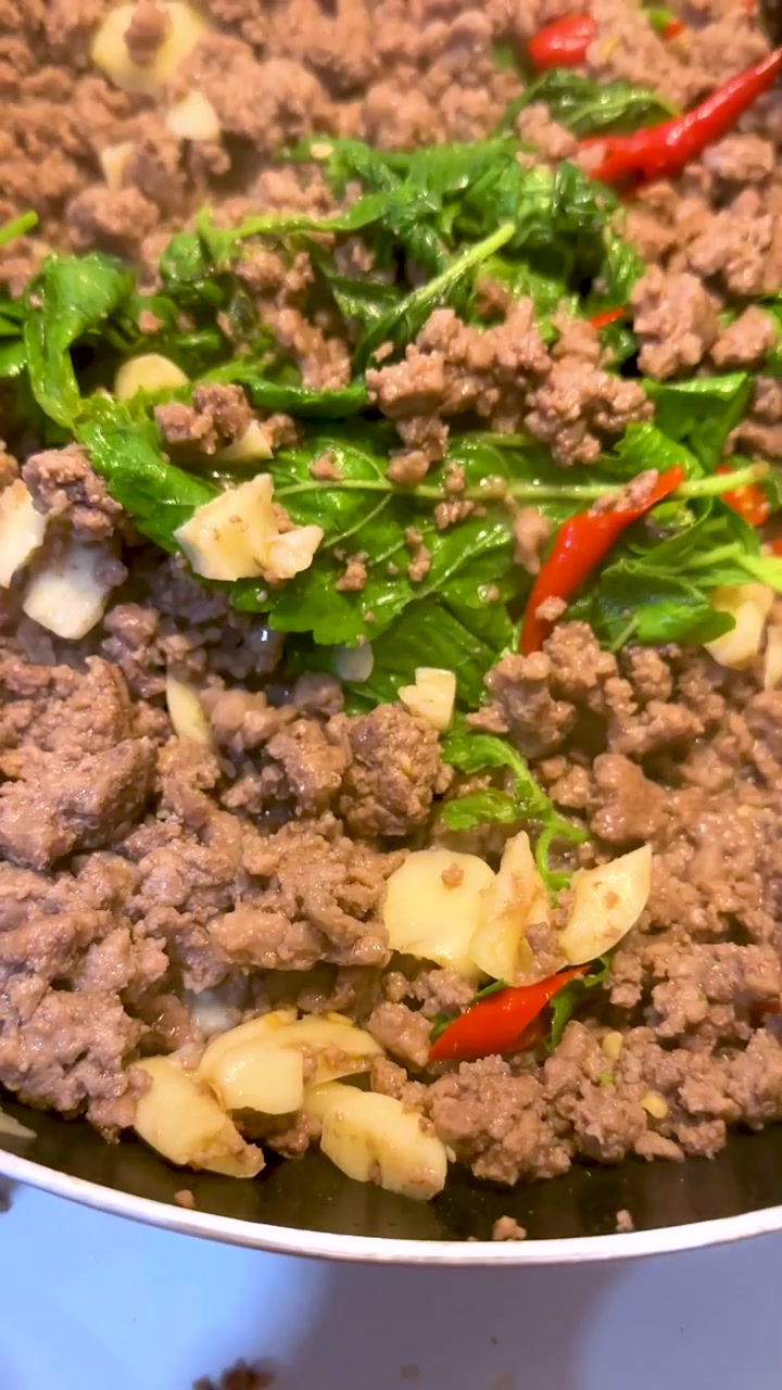 Sebuah spatula kayu mengaduk daging babi cincang, kemangi suci, dan cabai merah dalam wajan panas.