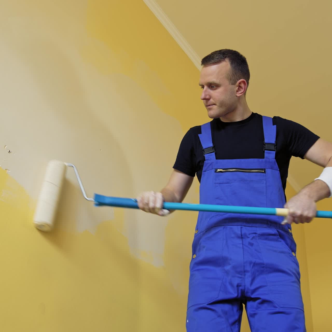 Builder painting wall in the room. Professional worker in workwear doing renovation in apartment with roller brush on a long stick. View from below