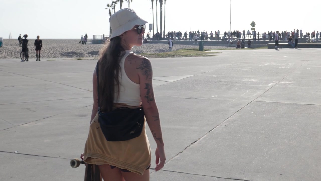 Kvinne fra Venice Beach, California, går på en skatepark-plass, med folkemengder, palmetrær og skateboardere i bakgrunnen. Hun har tatovert arm og bøttehatt, sett bakfra, et bilde av kystlivsstil.