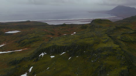 Aerial-view-of-mountains,-ocean-waves-crashing-to-shore-and-rugged-terrain-covered-with-grass-and-touches-of-snow-in-Iceland