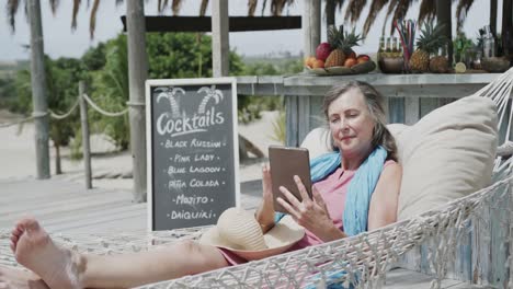 happy senior caucasian woman lying in hammock using tablet by beach bar, in slow motion