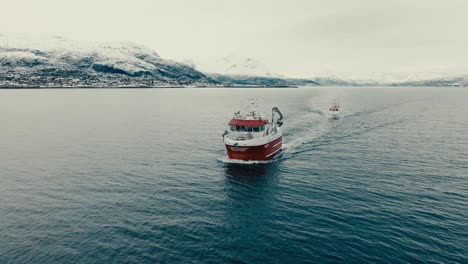 commercial fishing boat sailing through the ocean with snowy mountain in the distance in norway. - aerial shot