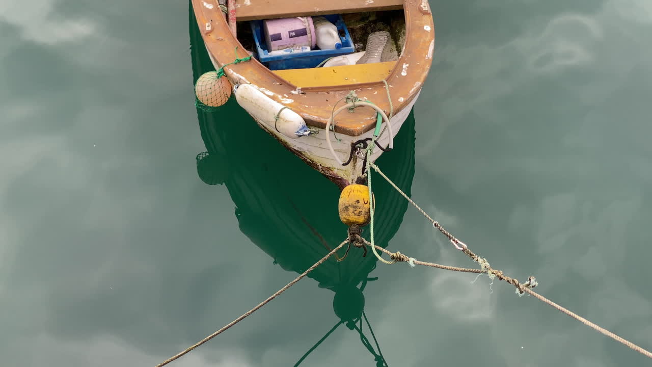 Weathered fishing boat floating on calm turquoise water, ropes and buoys reflected softly below for a tranquil harbor moment