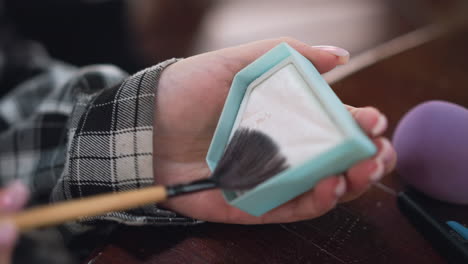 vista parcial de la mano de la dama sosteniendo el recipiente de polvo con cepillo frotando en polvo, aplicando maquillaje con herramientas de maquillaje esparcidas en la mesa, belleza y proceso de aseo personal en acción