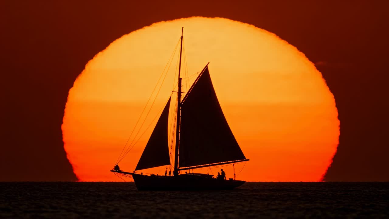 Vinden fylder sejlene på en tomastet sejlbåd, der glider henover det rolige åbne hav ved solnedgang, med besætning i bevægelse.