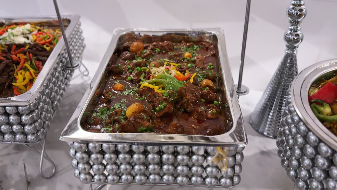 Buffet per banchetto al coperto con scaldavivande in acciaio inossidabile riempito con stufato di manzo brasato caldo, patate e contorno di verdure, servito su una linea di catering sotto la luminosa illuminazione della sala.