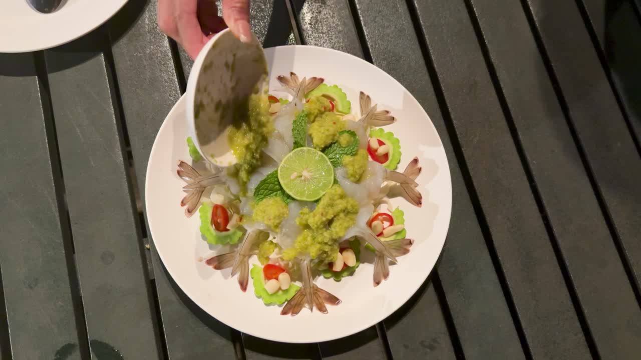 Hand pours green sauce onto raw shrimp seafood platter at night, overhead view, outdoor dining