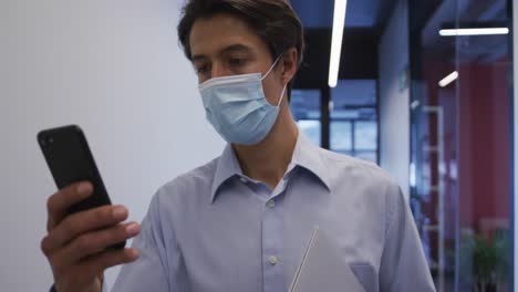 Caucasian-businessman-wearing-face-mask-walking-using-smartphone-in-office-corridor