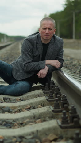 a man in a grey blazer and blue jeans lying on a railway track, looking contemplative and serious, the background features a rural landscape with trees, power poles, and a distant horizon