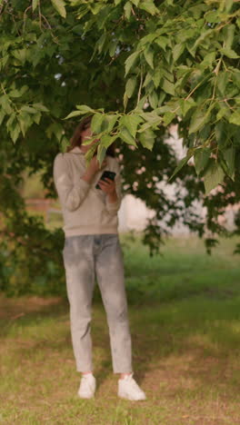 woman in casual attire and white sneakers stands under tree, focused on phone screen with hand touching hood collar, sunlight filtering through leaves casting soft reflection on her jeans