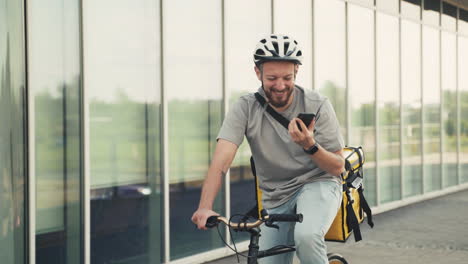 happy food delivery man wearing thermal backpack speaking on the phone on his bicycle