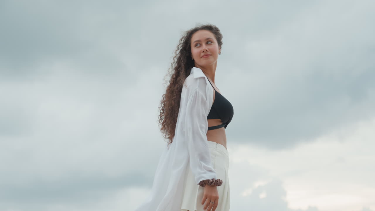 Donna sulla riva dell'isola che indossa una camicia bianca svolazzante, osserva il cielo nuvoloso con un paracadute ascensionale in lontananza, brezza calma, a piedi nudi, umore riflessivo, luce soffusa e coperta, movimento naturale dei capelli, postura rilassata
