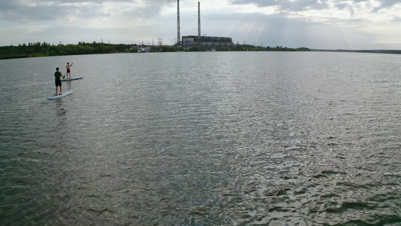 Young people sailing on water. Man and woman travelling on river on paddle boards on industry background in the evening. Summer rest.