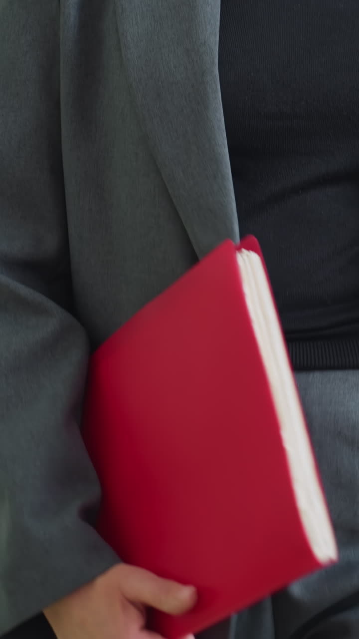 Businesswoman in gray suit holding red folder standing indoors ready for meeting or presentation with confident body language and professional attire in office or corporate environment