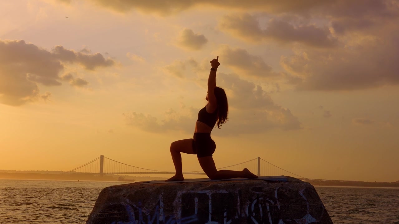 Donna che esegue la posa yoga dell'affondo basso durante il tramonto a New York. Silhouette di profilo di una donna su un tappetino da fitness su un molo di cemento. Nuvole dorate e ponte sospeso sullo sfondo