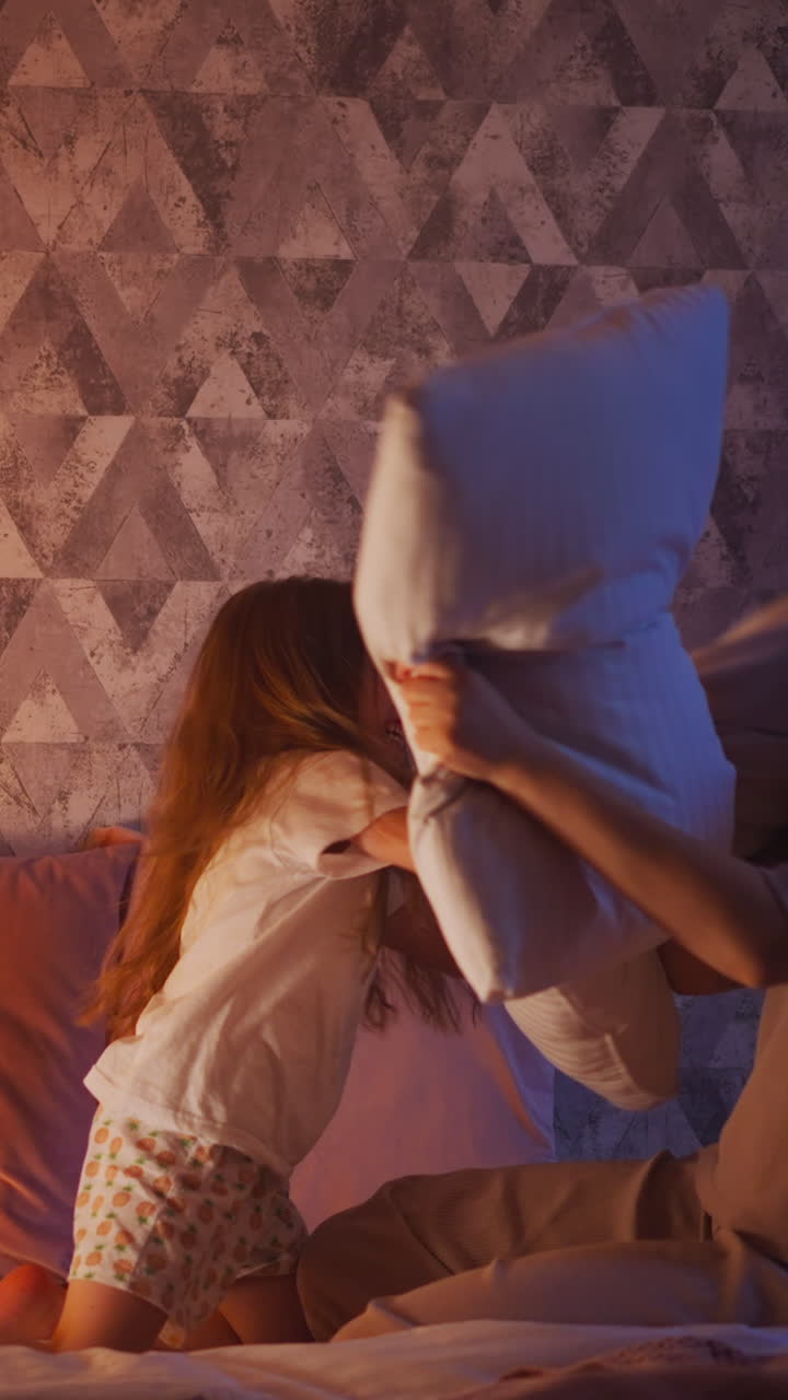 Funny little daughter and blonde mother in pajamas have pillow fight on large bed near play wigwam with glowing lamp inside in evening slow motion
