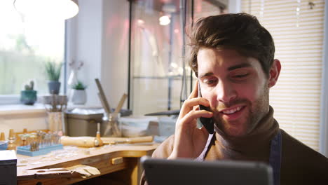 Male-Jeweller-Making-Phone-Call-Looking-At-Design-On-Digital-Tablet-In-Studio