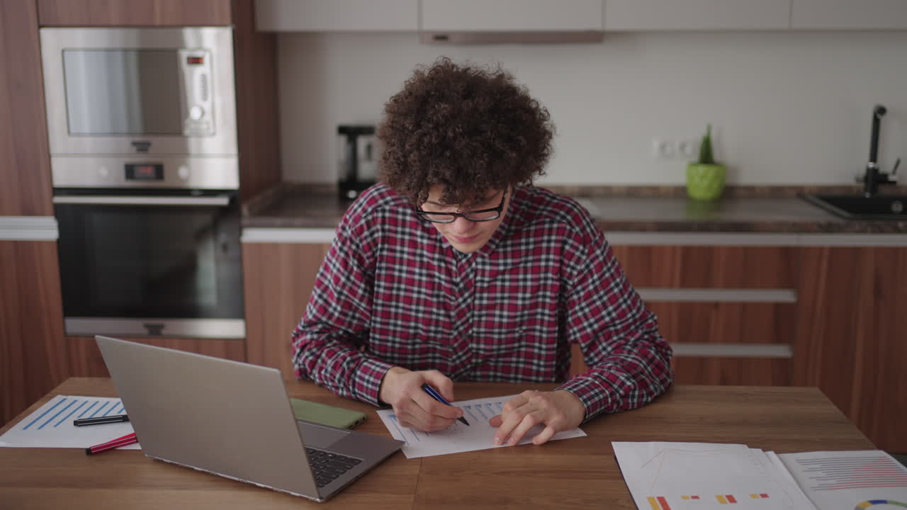 Curly haired Male student attractive young boy in glasses is studying at home using laptop typing writing in notebook. College student using laptop computer watching distance online learning seminar