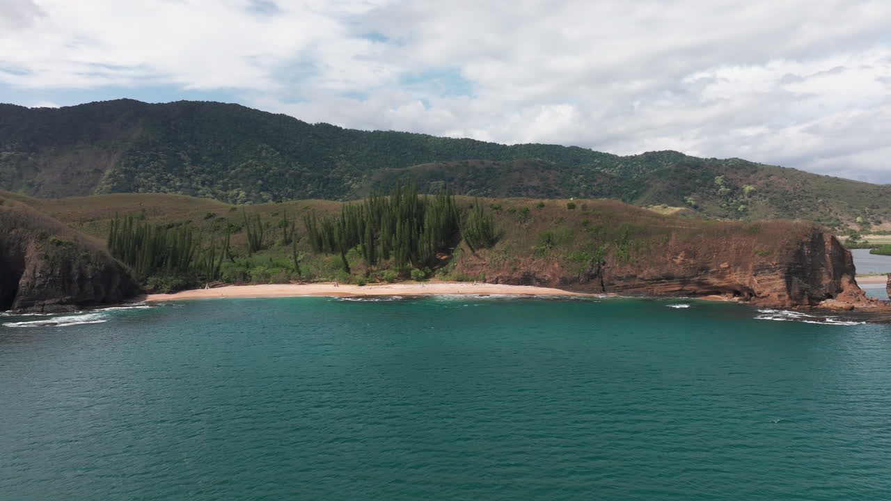 Drone view of Turtle Bay, New Caledonia, showing turquoise ocean waves washing onto a sandy beach surrounded by dramatic pine-covered hills