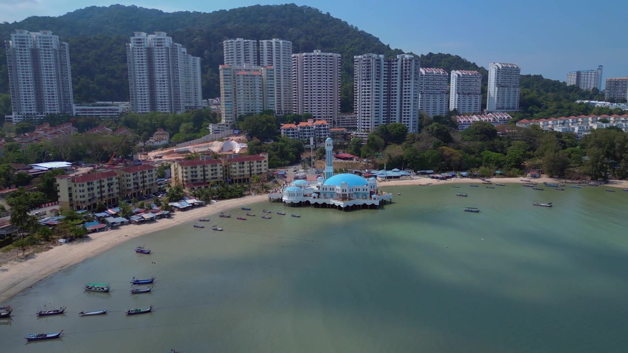 Moschea galleggiante di Tanjung Bungah, Penang, Malesia, con tradizionali barche da pesca lungo la costa ed edifici moderni sullo sfondo. Incredibile vista aerea panoramica da drone con ampia orbita.