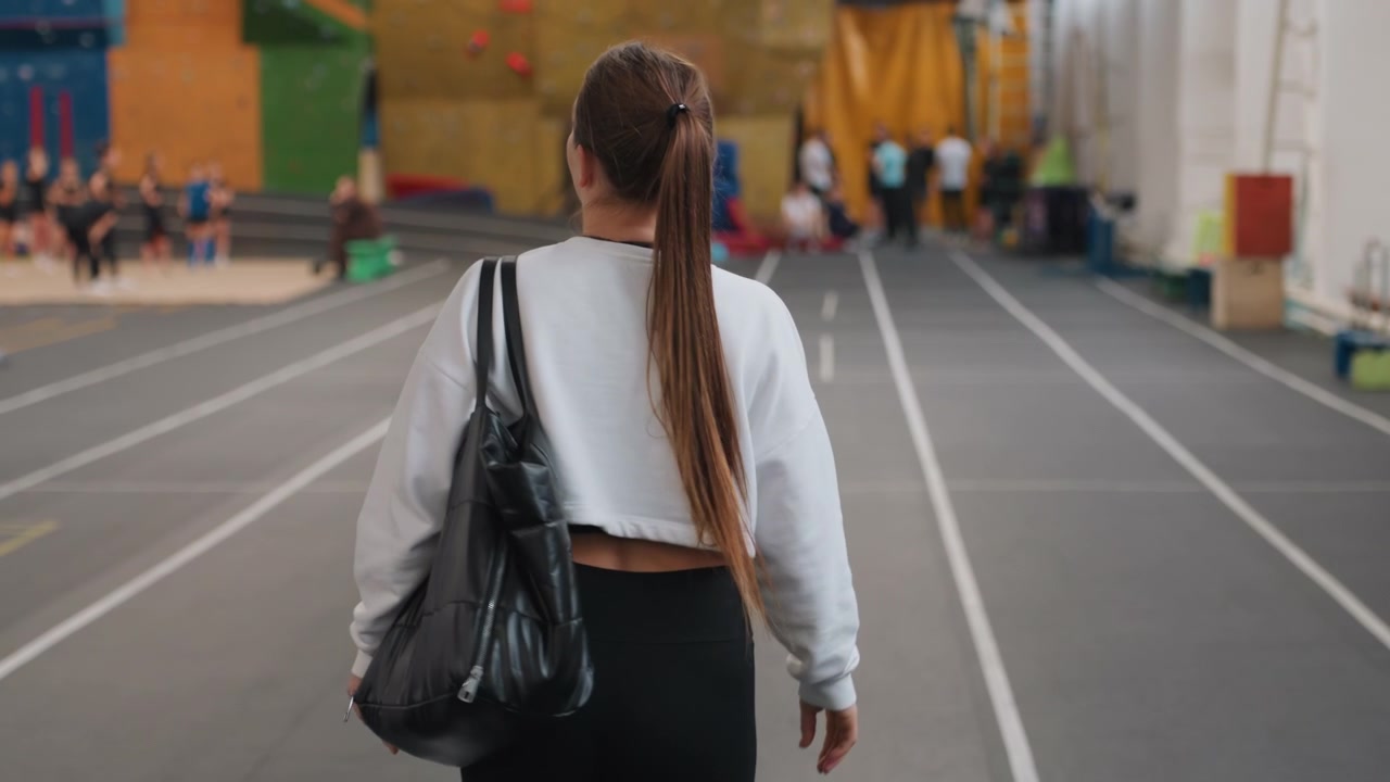 Atleta caucasica che cammina verso un campo affollato, coda di cavallo lunga e borsa lucida vista da dietro che mostra determinazione con spettatori sfocati e allestimento della competizione davanti, atmosfera da torneo e approccio concentrato.