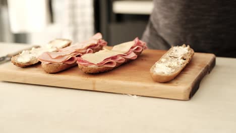 Caucasian-male-placing-sliced-Swiss-cheese-on-a-halved-brown-baguette-in-the-kitchen-on-the-countertop