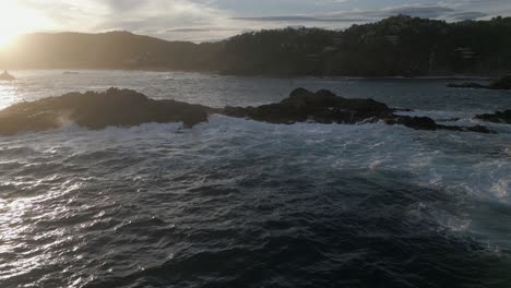 Golden-sunset-mist,-ocean-waves-crash-onto-rugged,-jagged-shore-rocks
