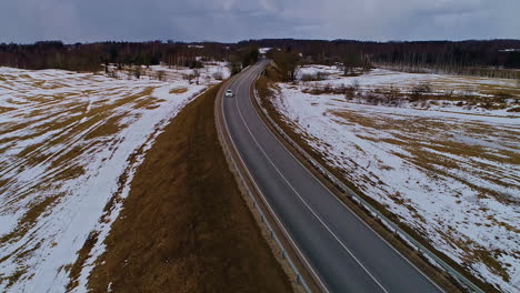 White-vehicle-driving-asphalt-road-with-snow-covered-fields,-aerial-drone-view