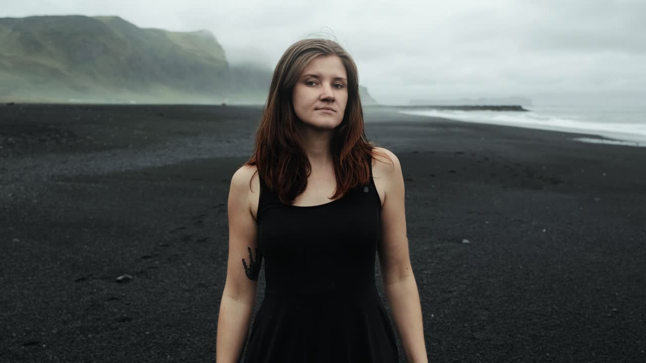 joven hermosa con vestido negro en la playa de arena negra de islandia, mirando a la cámara modelo posando, cámara lenta, dramáticas olas paisaje marino