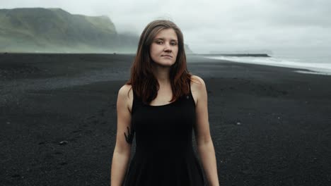 Joven-Hermosa-Mujer-Vestida-De-Negro-En-La-Playa-De-Arena-Negra-De-Islandia,-Mirando-A-La-Cámara-Modelo-Posando,-Cámara-Lenta,-Espectacular-Paisaje-Marino-De-Olas