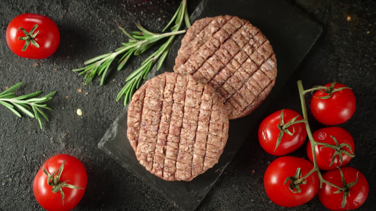 Raw burger on a board with rosemary and tomatoes rotates.