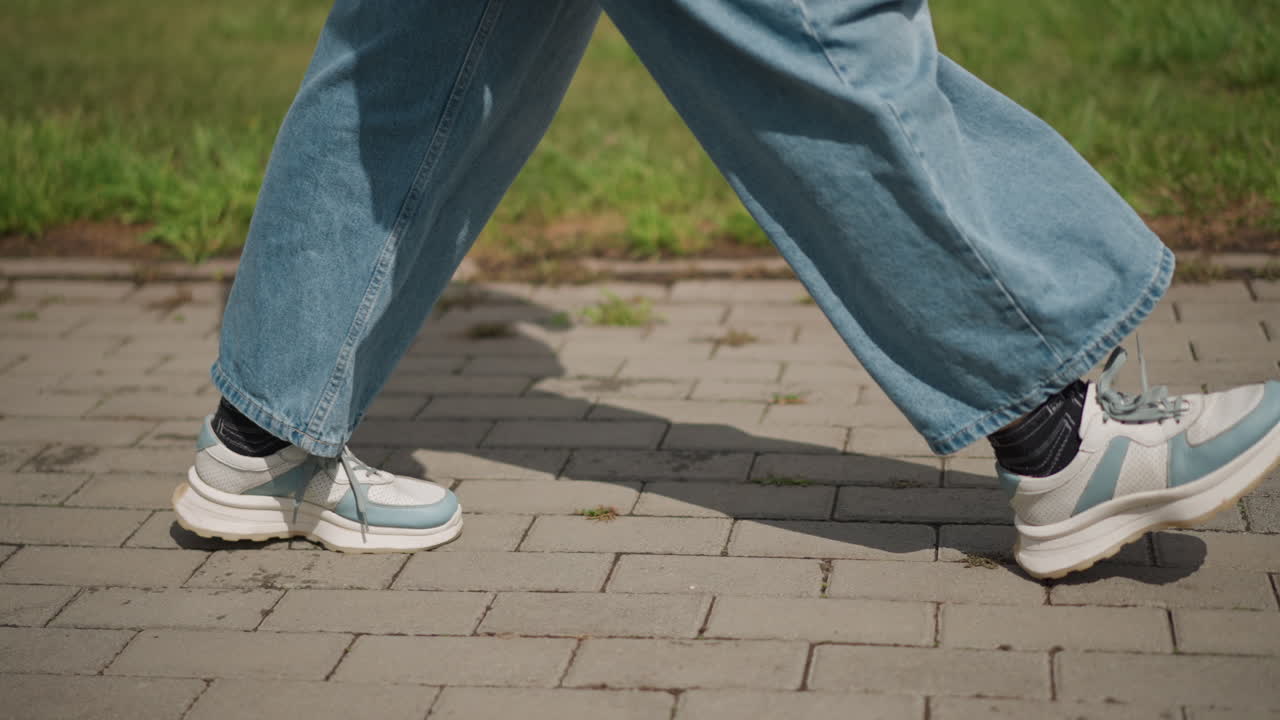 Primo piano di sneakers che camminano su un marciapiede in mattoni, ampi pantaloni di jeans che ondeggiano, inquadratura dal basso vicino a un bordo erboso, luce solare soffusa, passeggiata rilassata nel parco, attenzione al dettaglio della suola e dei lacci, atmosfera casual da streetwear