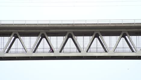 Two-bike-riding-men-driving-from-the-right-to-the-left-over-a-steel-construction-bridge-in-the-sky,-captured-from-lower-perspective