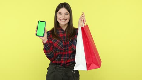 mujer positiva presumiendo con bolsas de compras sosteniendo un teléfono inteligente de pantalla verde