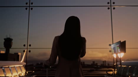 항공사 허브의 탑승 라운지에서 굴러가는 가방을 가진 아시아 사업가 여성의 뒷면, 스마트폰으로 이야기하고, 비행기를 기다리고, 공항 터미널에서 비행기가 창밖에서 이륙합니다.